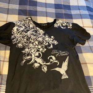 Affliction - Snygg svart t-shirt med ett detaljerat vitt blommönster och en fågel på framsidan. Perfekt för en stilren look med en touch av natur. Kortärmad och bekväm passform.