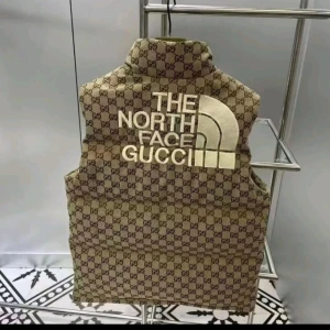Beige dunväst från The North Face x Gucci - Snygg beige dunväst med det ikoniska Gucci-mönstret och The North Face-logga. Västen har en hög krage och knappar framtill. Perfekt för att hålla stilen under kyliga dagar. Kan ta upp till 40dagar innan varan anländer