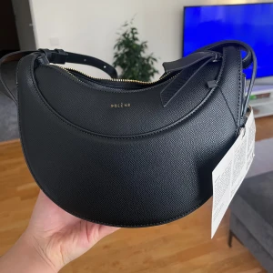 Svart axelväska från Polène - Snygg svart axelväska inspirerad av Polène i skinn med en elegant rundad form. Väskan har en justerbar axelrem och en praktisk dragkedja upptill. Perfekt för att bära dina dagliga nödvändigheter med stil.