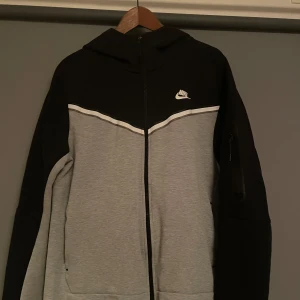 Grå och svart hoodie från Nike - Snygg grå och svart hoodie från Nike med dragkedja och huva. Den har en vit Nike-logga på bröstet och en praktisk ficka på ärmen. Den är i fint skick och stolek L.