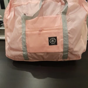 Rosa shoppingväska med grå handtag - Stor och praktisk rosa shoppingväska med grå handtag och dragkedja. Perfekt för resor eller shoppingturer. Väskan har en framficka med dragkedja för extra förvaring. Väskan kan du dra över ditt handtag på väskan så hänger den smidigt med, finns i svart,blå och rosa färg 