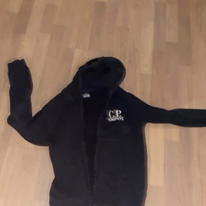 Svart huvtröja från C.P. Company - Snygg svart huvtröja från C.P. Company med unika glasögondetaljer på huvan. Perfekt för en cool och avslappnad stil. Tröjan har dragkedja och långa ärmar.