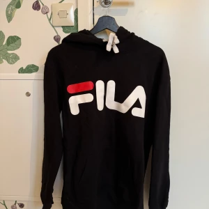Svart hoodie från FILA - Säljer en svart hoodie från FILA med stor logga i vitt och rött på framsidan. Tröjan har långa ärmar och en klassisk huva med dragsko. Perfekt för en avslappnad stil.
