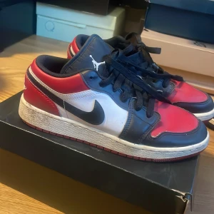 Air Jordan 1 Low  - Snygga Nike Air Jordans i klassisk färgkombination av rött, svart och vitt. Skorna har en ikonisk design med snörning och den välkända Air Jordan-loggan på hälen. Perfekta för sneakerheads som vill ha en tidlös stil.