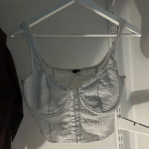 Vit broderad korsettopp - Säljer en stilren vit korsettopp med vackra broderade detaljer. Toppen har breda axelband och en figurnära passform som framhäver midjan. Perfekt för att skapa en elegant look.