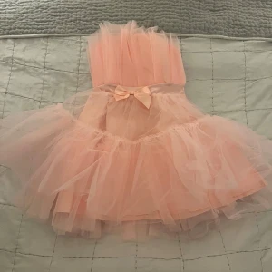 Rosa tyllklänning med volanger - Charmig rosa tyllklänning med volanger och en söt rosett i midjan. Perfekt för speciella tillfällen med sin fluffiga design och eleganta detaljer. ORGINAL PRIS: 600 kr