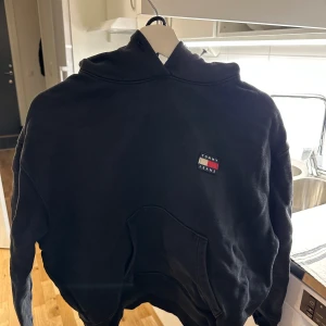 Svart hoodie från Tommy Hilfiger - Säljer en klassisk svart hoodie från Tommy Hilfiger med logotyp på bröstet. Tröjan har en stor ficka framtill och en bekväm huva. Perfekt för en avslappnad stil.