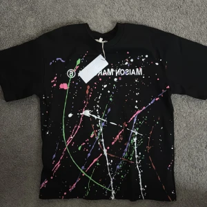 Svart t-shirt med färgstänk från Maison Margiela - Cool svart t-shirt från Maison Margiela med färgstänk i grönt, rosa, vitt och lila. Perfekt för en unik och konstnärlig stil. Kortärmad och i bomullsmaterial.