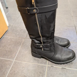 Svarta stövlar med dragkedja - Snygga svarta stövlar i skinn med dragkedja och dekorativa remmar. Perfekta för höst och vinter med en robust sula för extra grepp. Stövlarna har en klassisk design som passar till många olika stilar.