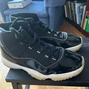 Svarta Nike Air Jordan 11 Retro - Snygga svarta Nike Air Jordans med hög design och glansiga detaljer. Skorna har snörning och en vit sula som kontrasterar mot det svarta. Perfekta för en sportig look.