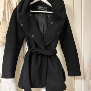 Svart kappa från NewYorker - Elegant svart kappa köpt på new Yorker för 699kr med knappar och knytskärp i midjan. Den har en stor huva och är perfekt för kyligare dagar. Stl 36 men passar även dig som är 34 möjligtvis 32. 