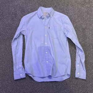 Blå skjorta från Lexington - Snygg blå skjorta från Lexington med klassisk krage och bröstficka. Skjortan har långa ärmar och knappar framtill. Perfekt för en stilren look.