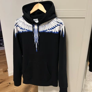 Marcelo Burlon tröja   - Snygg svart hoodie från Marcelo Burlon med ett iögonfallande vitt och blått vingmönster över axlarna. Perfekt för en cool och avslappnad stil. Den är i mycket bra skick. Storlek S men kan även passa dig som har M. Skriv för intresse!