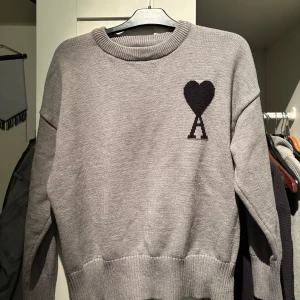 Grå stickad Ami Pari - Säljer en stilren grå stickad tröja med en svart hjärtdetalj på bröstet. Tröjan har långa ärmar och ribbstickade muddar vid ärmslut och nederkant. Perfekt för kyligare dagar.