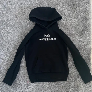 Svart hoodie från Peak Performance - Säljer en svart hoodie från Peak Performance med vit logga på bröstet. Tröjan har en klassisk design med huva och långa ärmar. Perfekt för en avslappnad stil.