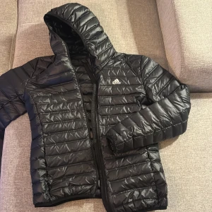 Svart dunjacka från Adidas - Snygg svart dunjacka från Adidas med huva och dragkedja. Perfekt för kyliga dagar med sitt quiltade mönster och sportiga look. Jackan har en diskret logga på bröstet och är lätt att matcha med olika outfits.