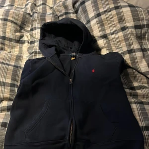 Mörkblå hoodie från Ralph Lauren - Säljer en snygg mörkblå hoodie från Ralph Lauren med dragkedja och en liten röd logga på bröstet. Perfekt för en avslappnad stil. Den har en klassisk passform med långa ärmar och en praktisk huva.