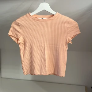 Persikofärgad ribbad topp från H&M - Säljer en  persikofärgad ribbad topp från H&M. Toppen har korta ärmar, volang kanter och  en rund halsringning. Är i storlek 134/140 