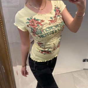 Gul t-shirt med blommönster - Snygg gul t-shirt med färgglatt blommönster och text på framsidan. Den har korta ärmar med volangdetaljer och en tight passform. Perfekt för en somrig look!