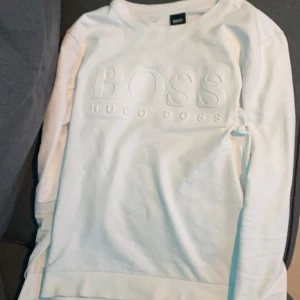 Vit tröja från Hugo Boss - Säljer en stilren vit tröja från Hugo Boss med deras logga i relief på framsidan. Tröjan har långa ärmar och en klassisk rund halsringning. Perfekt för en avslappnad och snygg look.