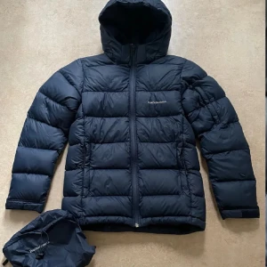 dunjacka från Peak Performance - Säljer en dunjacka från Peak Performance perfekt för kyliga dagar. Den är tillverkad i ett slitstarkt material och har en bekväm passform. Den har två små hål