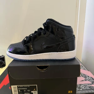 Jordan 1 Mid Space Jam - Snygga svarta Nike Air Jordans med glansig finish och vit sula. Skorna har klassisk snörning och en stilren design som passar perfekt för sneaker-entusiaster.