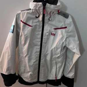 Vit vindjacka från Helly Hansen - Säljer en vit vindjacka från Helly Hansen med röda detaljer och dragkedja. Jackan har en hög krage och justerbar huva. Perfekt för blåsiga dagar och utomhusaktiviteter.