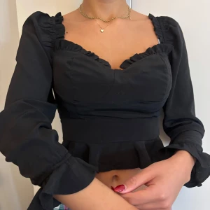  (off-shoulder) blus med volangdetaljer - Snygg svart blus med volangdetaljer och långa ärmar. Blusen har en sweetheart-neckline design och en croppad passform. Sitter ganska croppat på mig då jag är lång. Helt ny bara haft ett tag i garderoben.