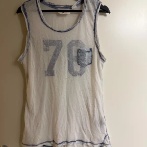 Vit ärmlös topp från H&M - Säljer en vit ärmlös topp från H&M med blå detaljer och ett stort tryck av siffran 70 på framsidan. Toppen har en liten bröstficka och är perfekt för en avslappnad stil.