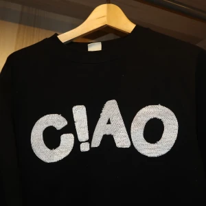 Svart tröja med 'CIAO' tryck - Snygg svart tröja med ett stort vitt 'CIAO' tryck på framsidan. Perfekt för en avslappnad stil. Tröjan har långa ärmar och en rund halsringning.