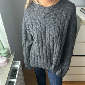 Grå stickad tröja - Mysig grå stickad tröja med kabelstickat mönster. Tröjan har långa ärmar och ribbade muddar vid ärmslut och nederkant. Perfekt för kyliga dagar.