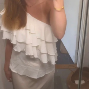 Vit oneshoulder topp med volanger - Snygg vit oneshoulder topp med flera lager volanger. Perfekt för en somrig look eller festliga tillfällen. Toppen har en luftig passform och är tillverkad i ett lätt material.