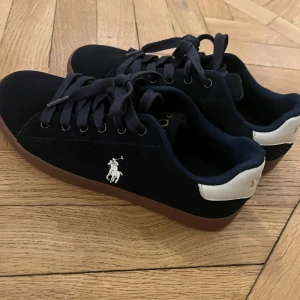 Marinblå  sneakers från Polo Ralph Lauren - Snygga marinblå  sneakers med bruna sulor från Polo Ralph Lauren. Skorna har en vit logga på sidan och texten 'POLO' i guld på hälen. De är utrustade med svarta snören och har en klassisk design.