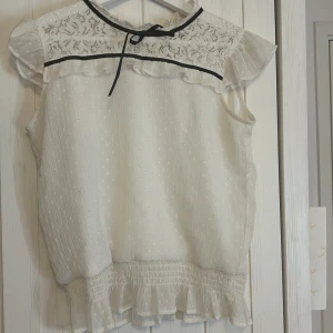 Vit spetsblus från Lindex - Söt vit blus från Lindex med spetsdetaljer och volangkant. Blusen har en knytning vid halsen och är ärmlös med ett lätt prickigt mönster. Perfekt för en somrig look!🤍 Blusen är i gott skick!!🤍