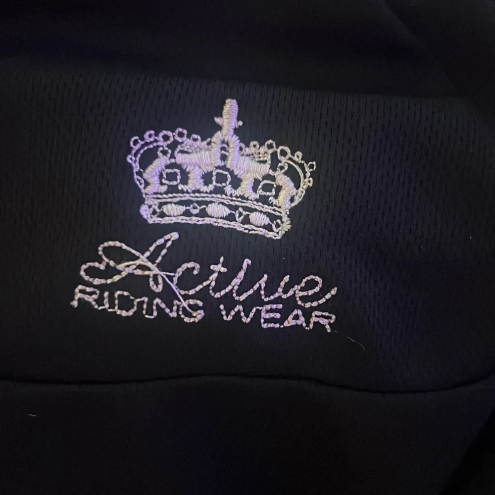 Snygg svart tröja från Active Riding Wear med broderad krona och text på bröstet. Perfekt för ridning och andra utomhusaktiviteter. Tröjan har en dragkedja framtill och en hög krage för extra skydd mot väder och vind. Väldigt tunn så perfekt att ha under en jacka. Neuletakit & Villapaidat.