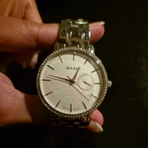 Silverfärgad klocka från GANT - Elegant silverfärgad klocka från GANT med en stilren urtavla och glittrande detaljer runt boetten. Perfekt för att ge en touch av lyx till din stil. Armbandet är i metall och matchar den klassiska designen.