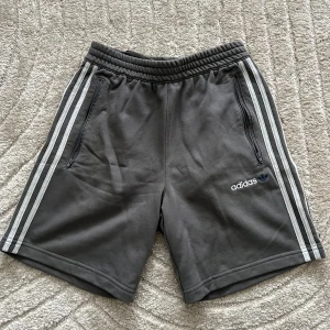 Grå shorts från Adidas - Snygga grå shorts från Adidas med klassiska vita ränder längs sidorna. De har en elastisk midja och praktiska dragkedjefickor. Perfekta för en sportig look!