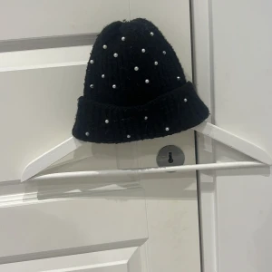 Svart stickad mössa med pärlor - Snygg svart stickad mössa dekorerad med vita pärlor. Den är lite nopprig men annars i bra skick. Den är i storlek 7-9 år gammal som är storlek 52/53