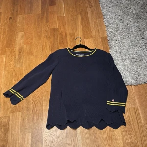 Mörkblå blus med gula detaljer - Snygg mörkblå blus med långa ärmar och gula ränder vid ärmslut och hals. Blusen har en vågig nederkant som ger en unik touch. Perfekt för en stilren look.