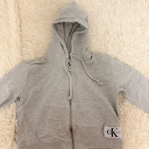 Grå hoodie från Calvin Klein - Säljer en stilren grå hoodie från Calvin Klein med dragkedja och justerbar huva. Perfekt för en avslappnad look. Den har en logga på framsidan och är tillverkad i mjukt material.