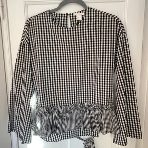 Svartvit rutig blus med volang - Säljer en svartvit rutig blus från Zara med volangdetalj i nederkant. Blusen har långa ärmar och knytband i ryggen för en fin passform. Perfekt för en stilren look!