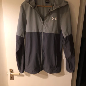 Grå vindjacka från Under Armour - Snygg grå vindjacka från Under Armour med dragkedja och luva. Perfekt för blåsiga dagar. Jackan har en stilren design med två nyanser av grått och en diskret logga på bröstet.