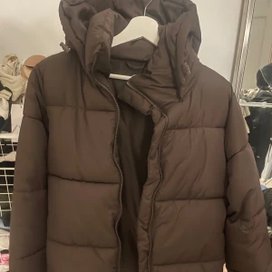 Brun dunjacka med huva  - Säljer en brun dunjacka med huva, perfekt för kyliga dagar. Jackan har en dragkedja framtill och är quiltad för extra värme. Från cubus