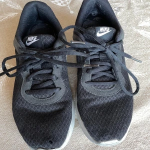 Svarta Nike Tanjun träningsskor  - Säljer ett par svarta Nike sneakers (äkta såklart, köpta på stadium) med vit sula och klassisk swoosh-logga. Skorna har snörning och har meshtyg. Perfekta för vardagsbruk och träning. Använda men i gott skick med några skavanker (se bild). Storlek 38. 