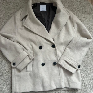 Beige ullkappa från MNG - Snygg beige kappa i ull från MNG med dubbelknäppning och stora svarta knappar. Perfekt för kyligare dagar. Nypris 1049kr