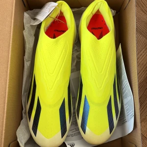 Adidas X Crazyfast Elite LL FG Fotbollsskor - Säljer ett par Adidas X Crazyfast Elite LL FG fotbollsskor i en iögonfallande gul färg med svarta och blå detaljer. Skorna har en strömlinjeformad design utan snörning och är perfekta för spel på fasta underlag.