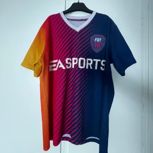 Flerfärgad fotbollströja från EA Sports - Snygg och unik fotbollströja från EA Sports med korta ärmar. Tröjan har en cool design i blått, rött och gult med ett stort '18' på ryggen och 'FUT 18' på bröstet. Perfekt för gamingfans och fotbollsälskare! Lite färg på märket har försvunnit och några fläckar.