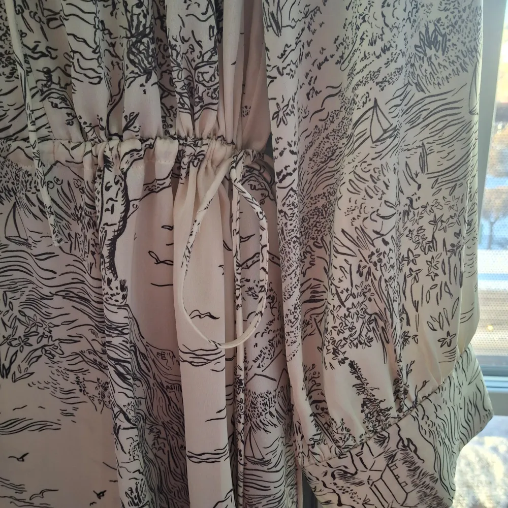 Säljer en elegant beige klänning från H&M med ett unikt svart mönster. Klänningen har långa ärmar och ett knytband i midjan för en smickrande passform. Perfekt för både vardag och speciella tillfällen.. Mekot.