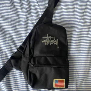 Svart axelväska från Stüssy  - Snygg svart axelväska från Stussy med justerbar rem och dragkedja. Väskan har en cool logga framtill och en patch med amerikanska flaggan. Perfekt för att bära dina essentials med stil. Den har också väldigt mycket plats den får plats med ett par skor 
