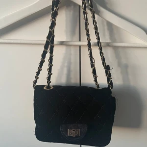Svart quiltad axelväska med kedja - Snygg svart axelväska med quiltat mönster och kedjedetaljer. Väskan har en praktisk storlek och stängs med ett metallspänne framtill. Perfekt för att ge en stilren touch till din outfit.